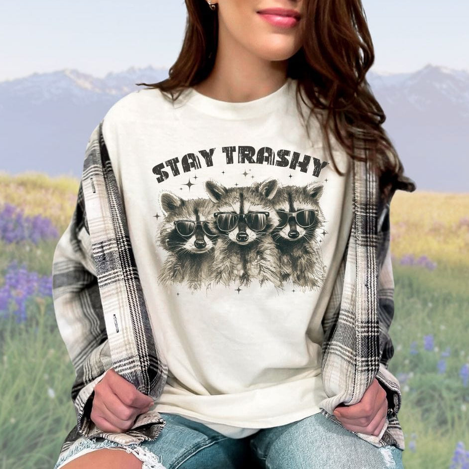 A person wearing an ivory crew neck t-shirt with the phrase 'STAY TRASHY' printed above an illustration of three raccoons in sunglasses.
