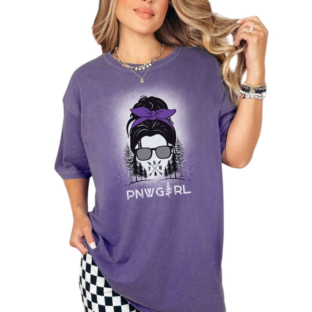 A woman wearing a purple vintage style t-shirt with a graphic print of a girl's face and the text 'PNWGIRL'. The shirt is round-necked, and the woman is accessorized with a necklace and sunglasses.
