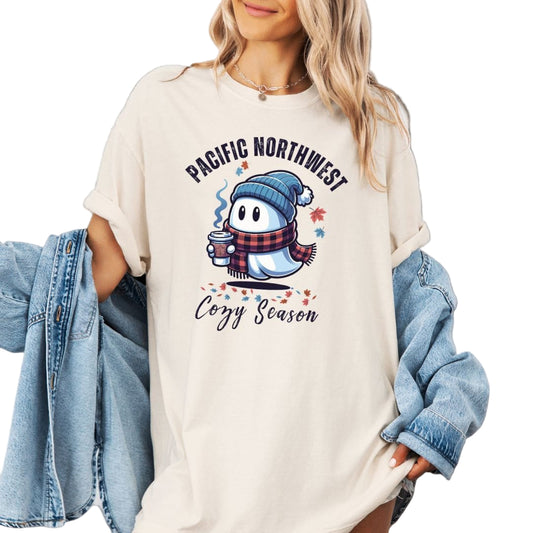 Person wearing a white sweatshirt with a Pacific Northwest Cozy Season design, holding a denim jacket.