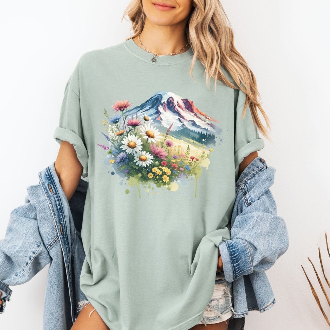 Woman wearing a sage green Comfort Colors t-shirt featuring a watercolor graphic print of Mount Rainier surrounded by a vibrant meadow of colorful wildflowers and daisies.