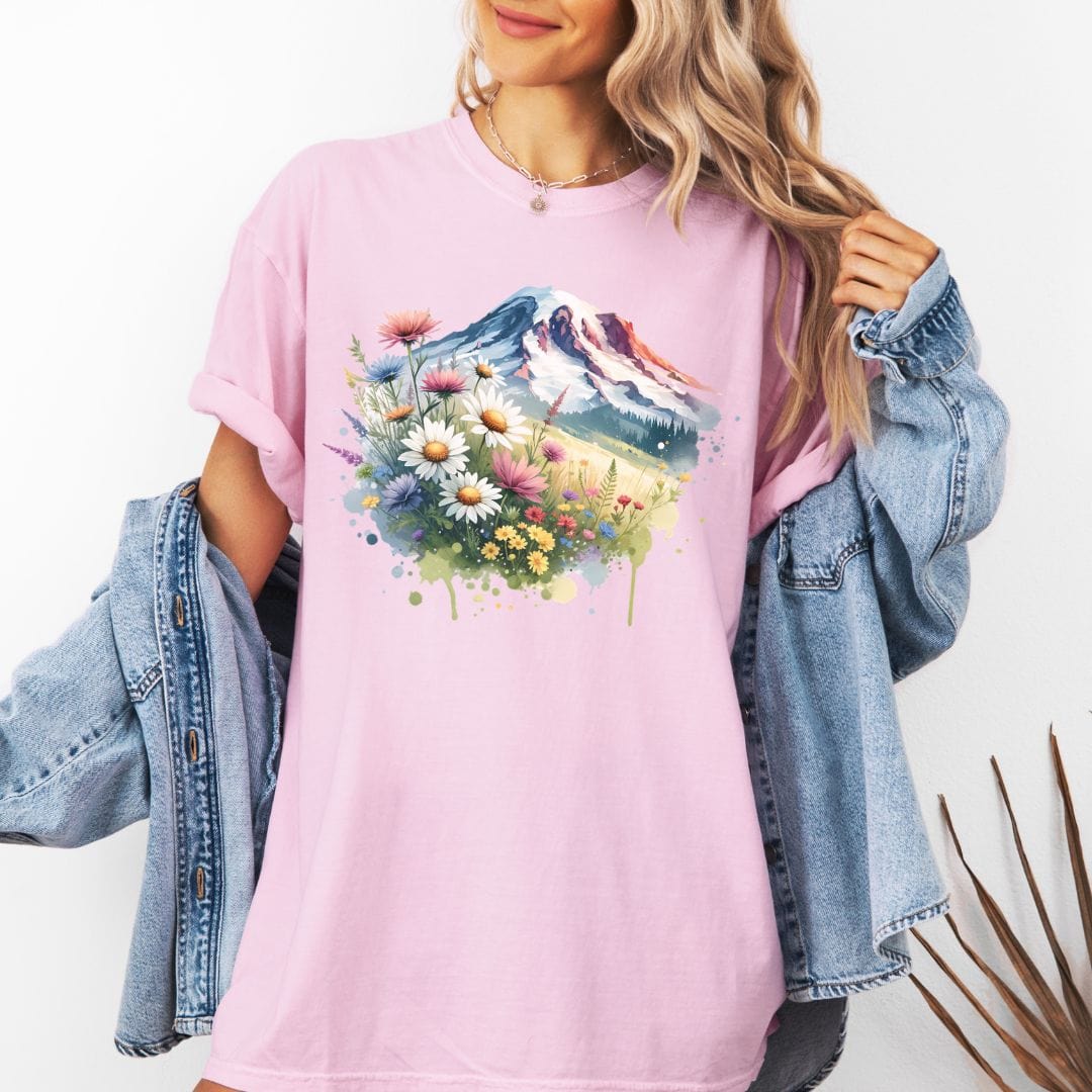Woman wearing a pink Comfort Colors t-shirt featuring a watercolor graphic print of Mount Rainier surrounded by a vibrant meadow of colorful wildflowers and daisies.
