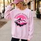A woman wearing a light pink crewneck sweatshirt with a colorful 'MOUNT RAINIER' mountain graphic on the front, paired with a matching pink cap, standing on a sidewalk.