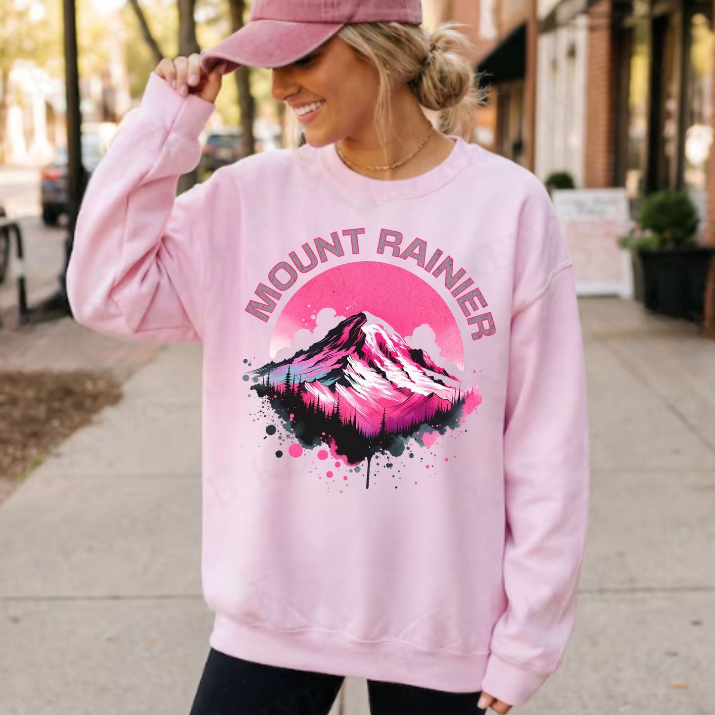 A woman wearing a light pink crewneck sweatshirt with a colorful 'MOUNT RAINIER' mountain graphic on the front, paired with a matching pink cap, standing on a sidewalk.