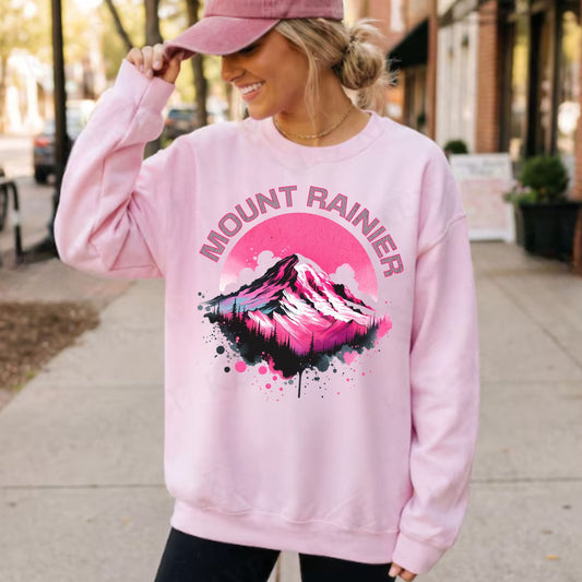 A woman wearing a light pink crewneck sweatshirt with a colorful 'MOUNT RAINIER' mountain graphic on the front, paired with a matching pink cap, standing on a sidewalk.