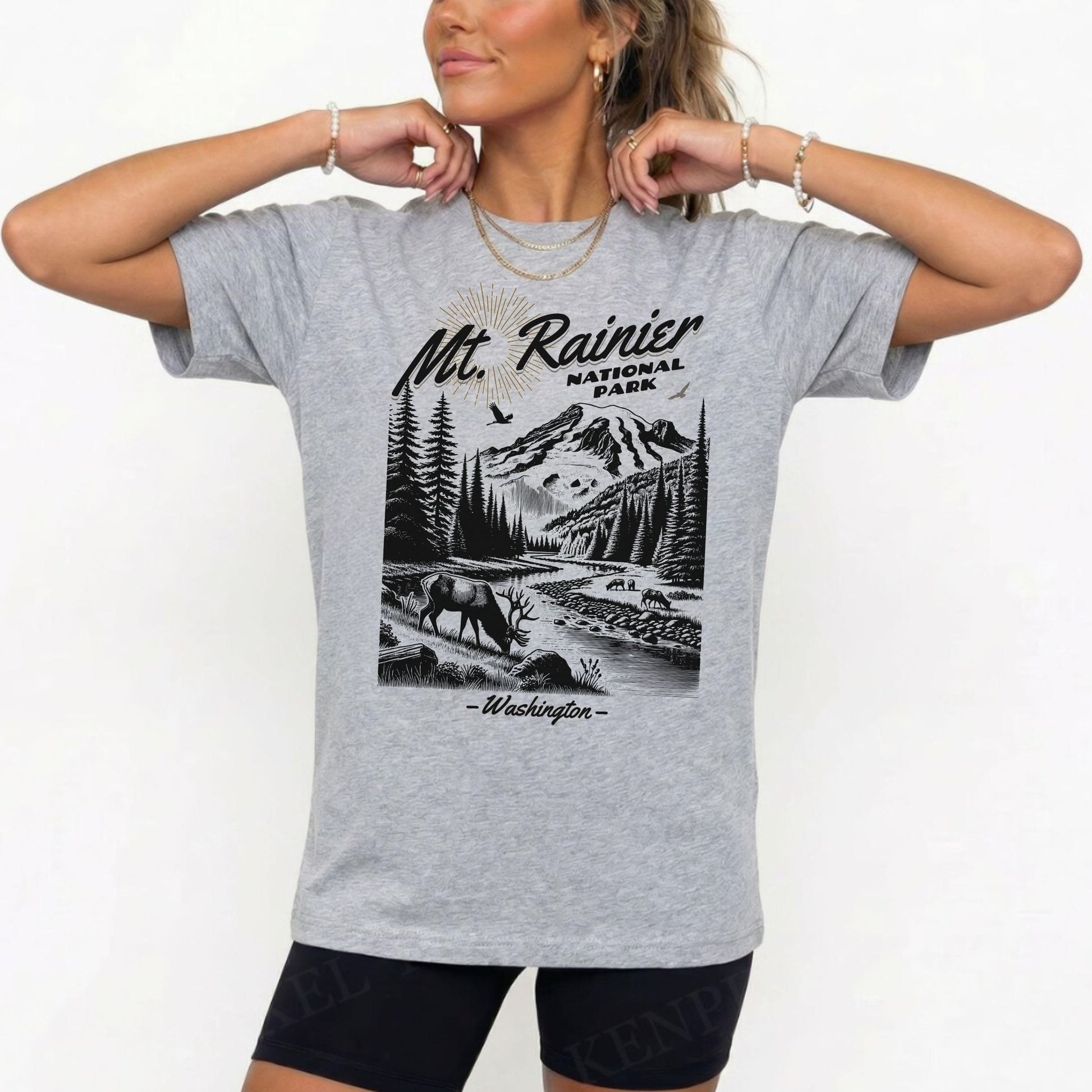 A woman wearing a heather gray graphic t-shirt featuring a black and white illustration of Mt. Rainier National Park, Washington, paired with black shorts against a plain white background.