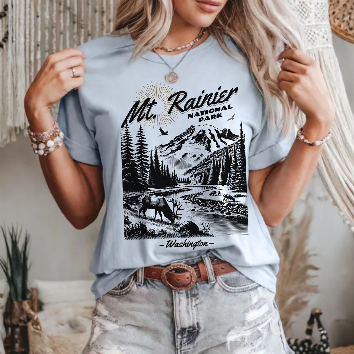 A woman wearing a light blue graphic t-shirt featuring a black and white illustration of Mt. Rainier National Park, paired with distressed denim shorts in a cozy boho-style room.