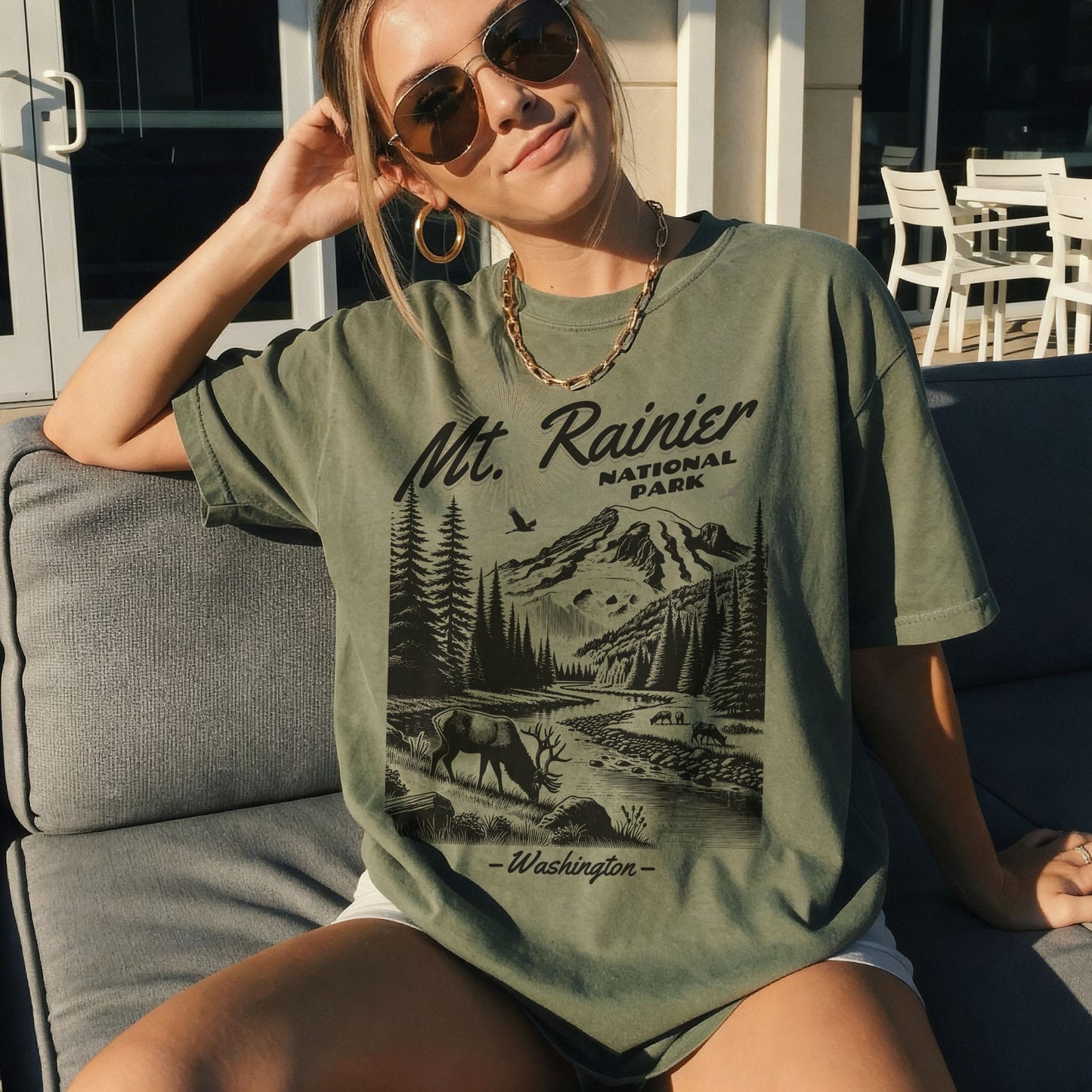 A woman wearing a military green graphic t-shirt with a black and white print of Mt. Rainier National Park, sitting on a gray outdoor couch.