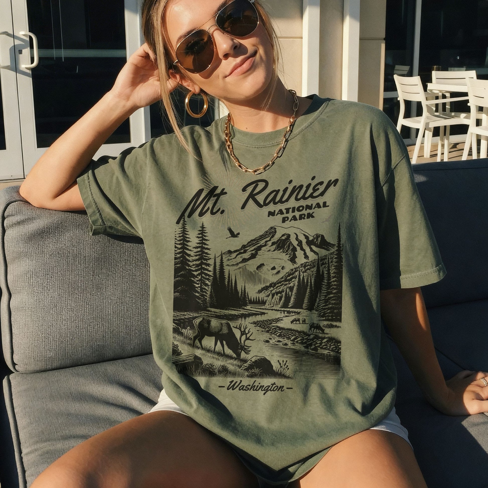 A woman wearing a military green graphic t-shirt with a black and white print of Mt. Rainier National Park, sitting on a gray outdoor couch.
