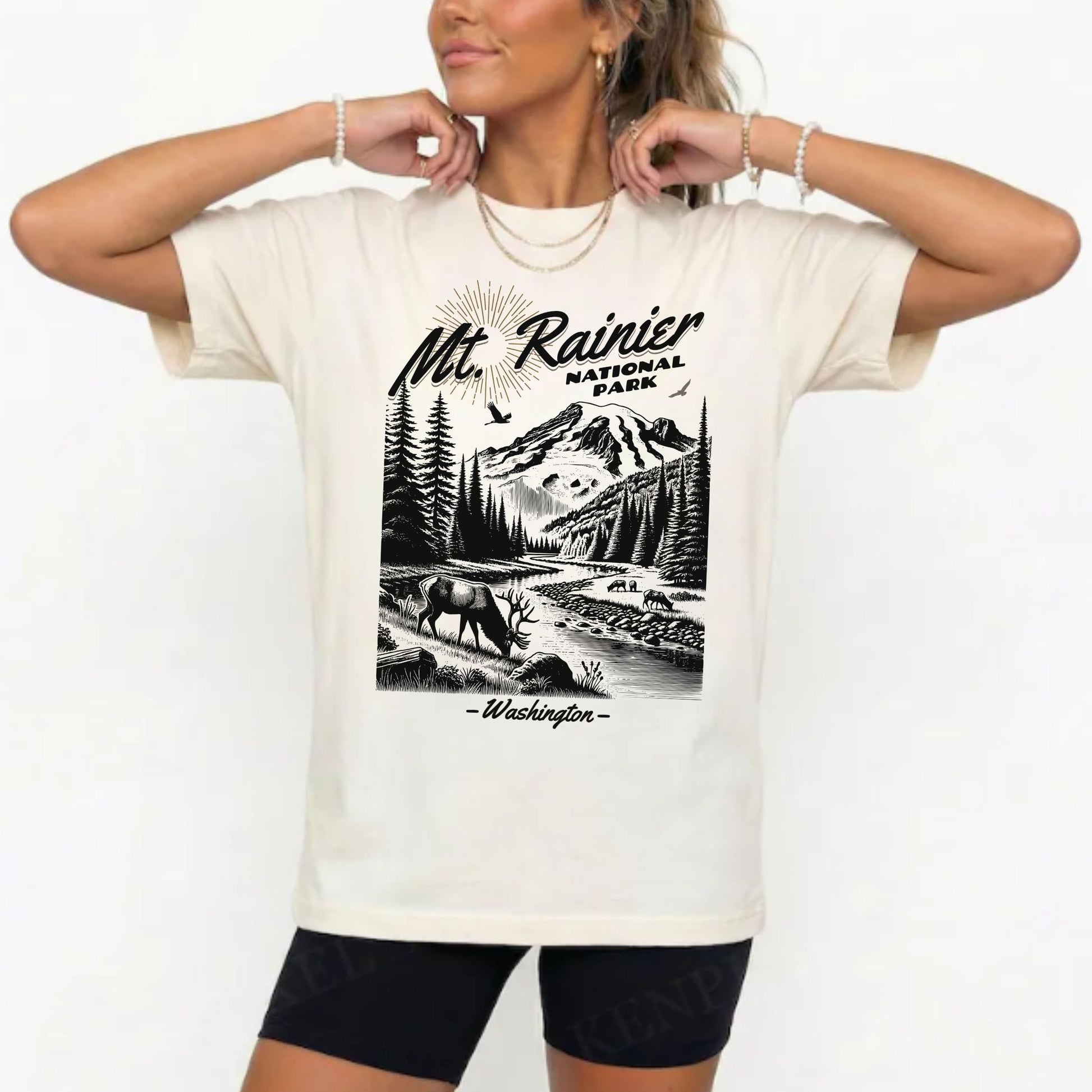 A woman wearing a cream-colored t-shirt with a black graphic print of Mt. Rainier National Park, paired with black bike shorts, against a plain white background.