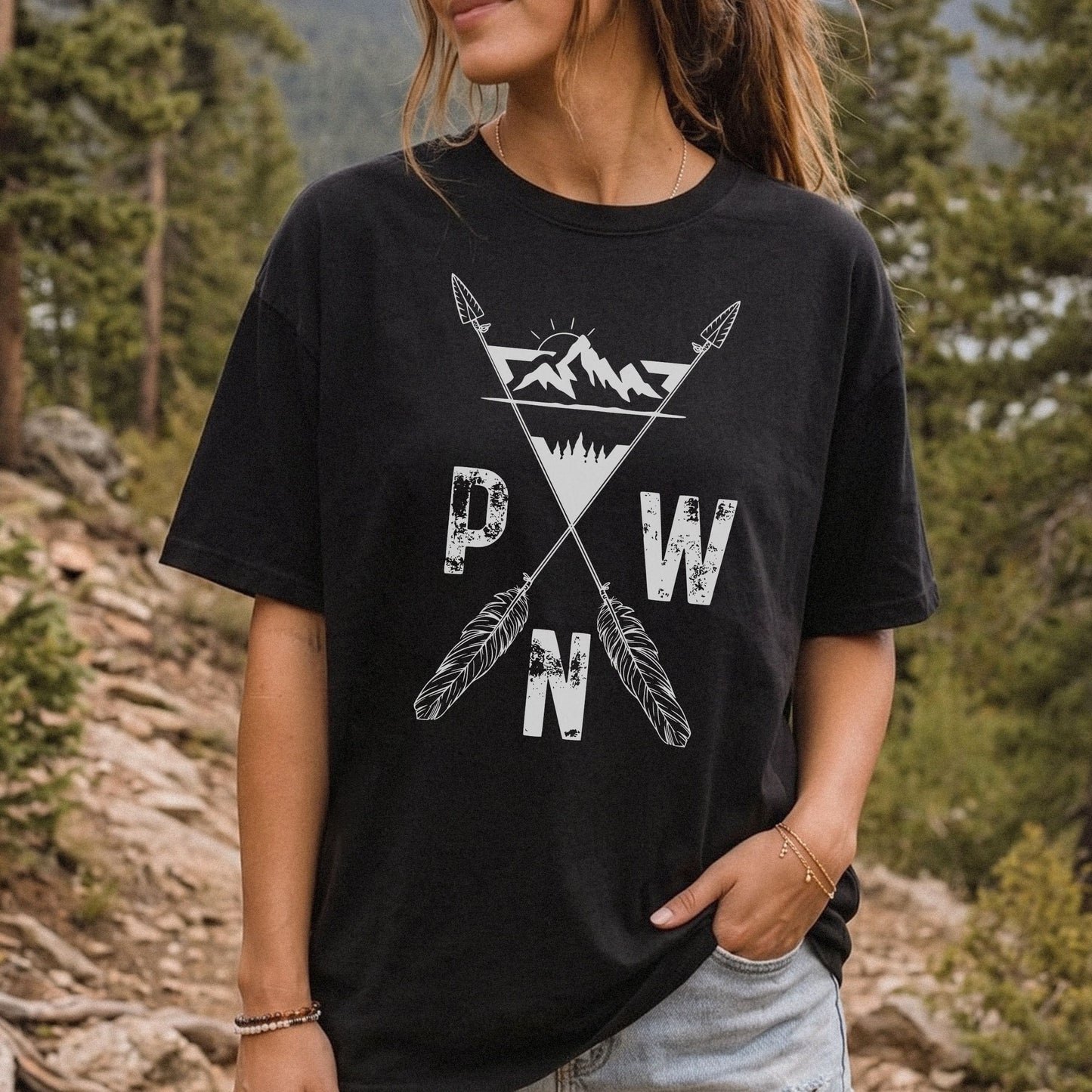 A woman wearing a black graphic t-shirt with a white distressed design featuring crossed arrows, a mountain triangle, and the letters 'PWN', photographed outdoors in a forest setting.
