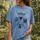 A woman wearing a light blue graphic t-shirt with a black mountain and arrow design and the letters 'PWN' on the front, standing in a forest setting.