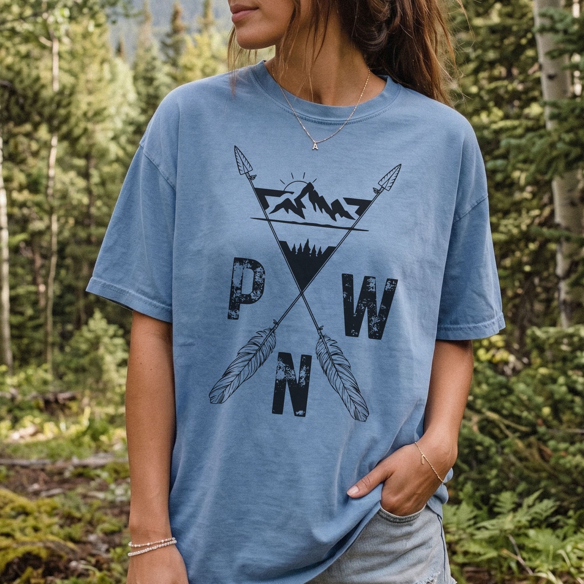 A woman wearing a light blue graphic t-shirt with a black mountain and arrow design and the letters 'PWN' on the front, standing in a forest setting.