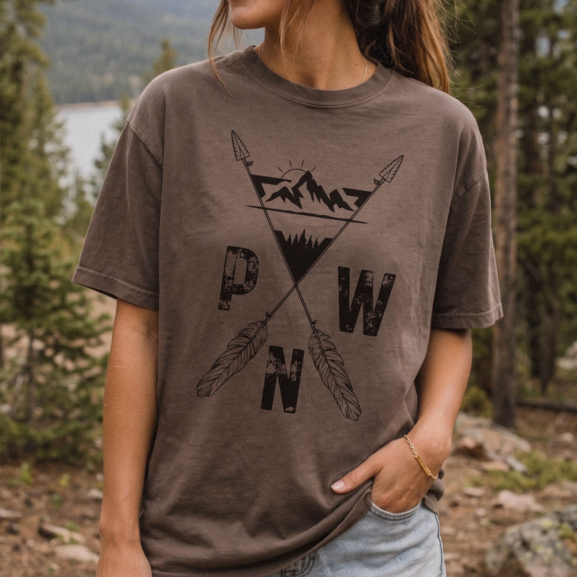 A woman wearing a brown short-sleeve graphic t-shirt with a black mountain and crossed arrows design, standing outdoors in a forest setting.