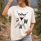 A woman wearing a cream-colored t-shirt with a black graphic of crossed arrows, mountains, and the letters 'PWN', set against a blurred outdoor background of trees and a lake.