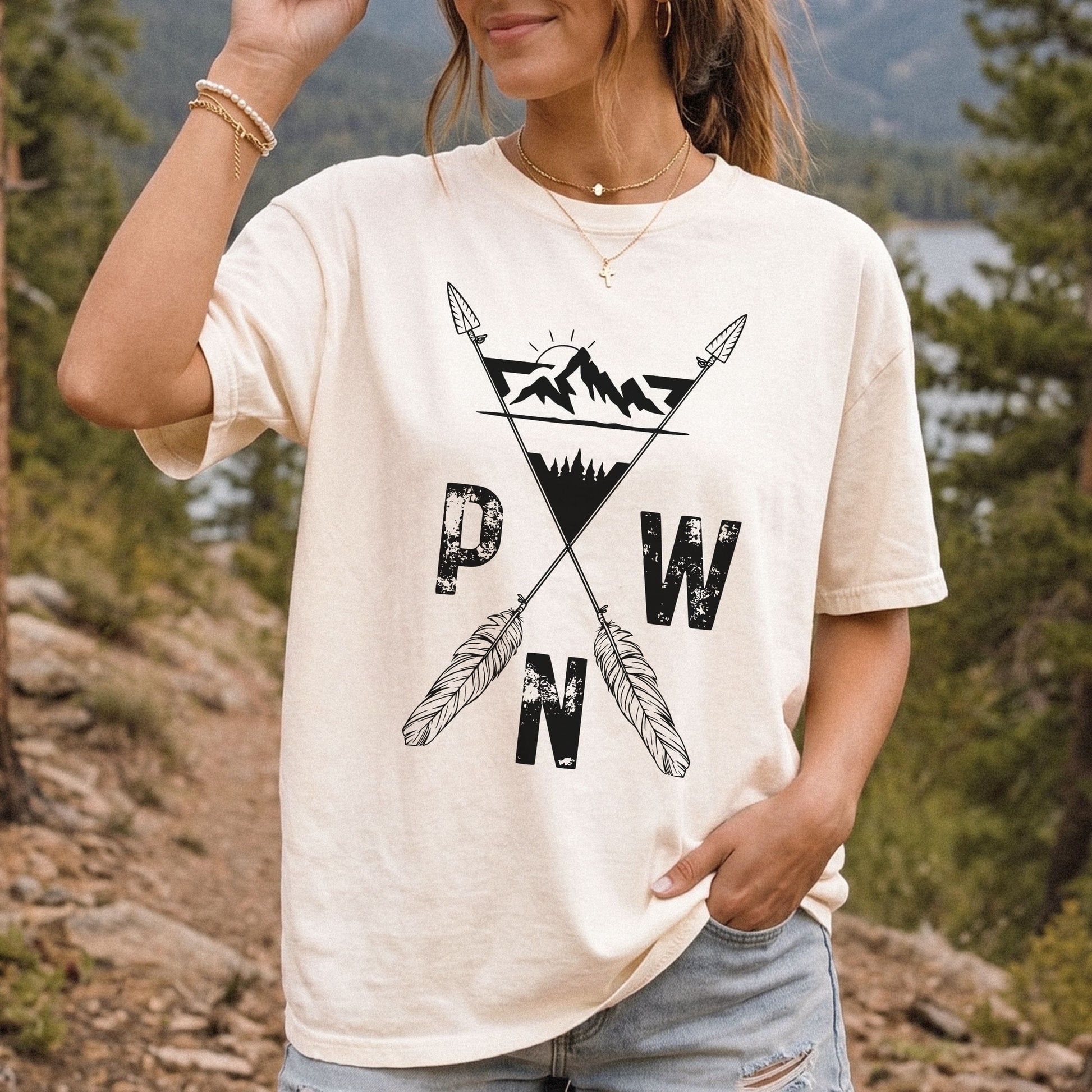 A woman wearing a cream-colored t-shirt with a black graphic of crossed arrows, mountains, and the letters 'PWN', set against a blurred outdoor background of trees and a lake.