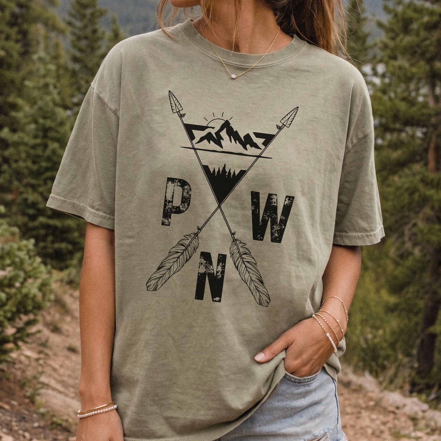 A woman wearing a khaki graphic t-shirt with a black mountain and feather design, standing outdoors in a forest setting.