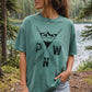 A woman wearing a sage green comfort colors t-shirt with a black mountain and arrow design and the letters 'P W N' on the front, standing outdoors in front of a forest lake.