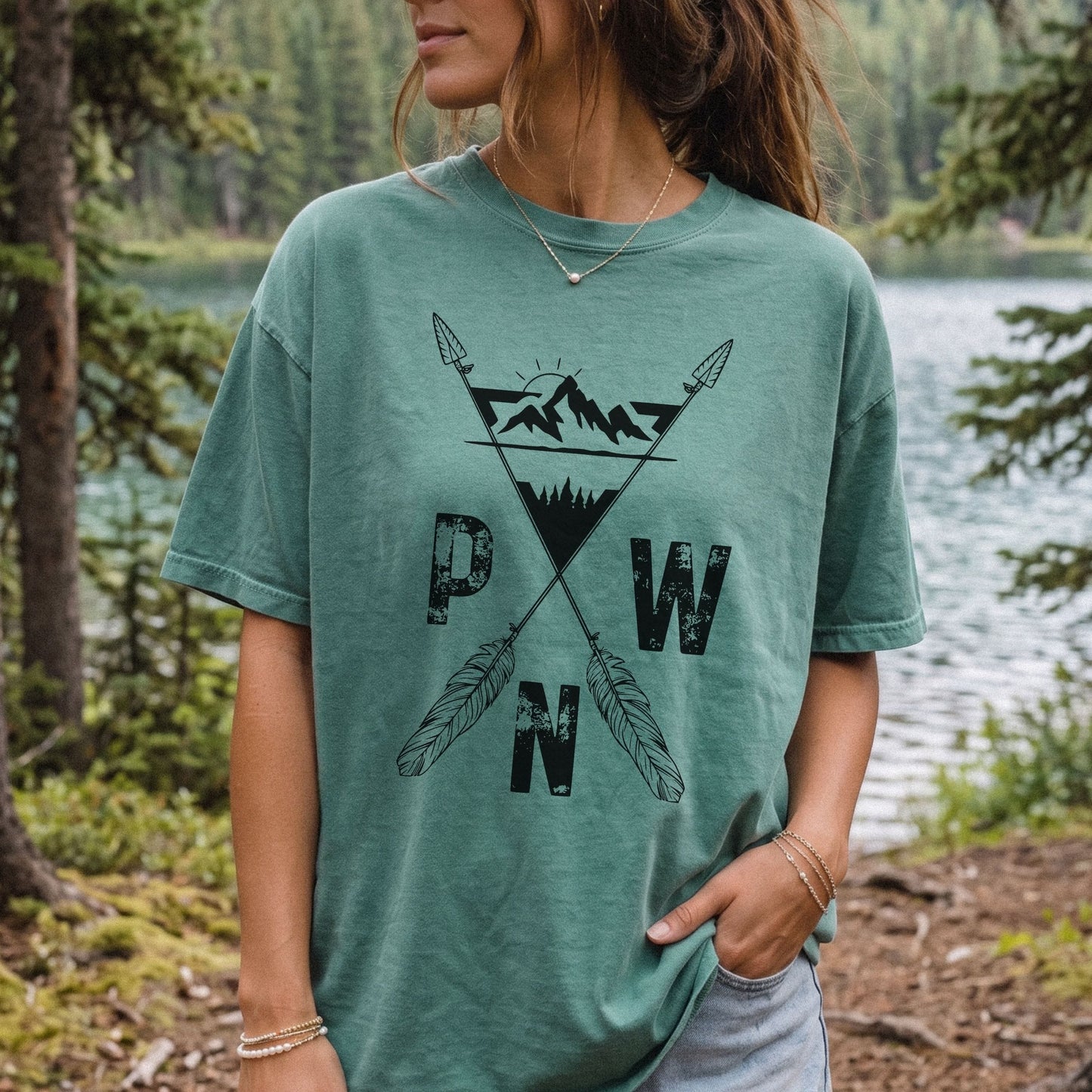 A woman wearing a sage green comfort colors t-shirt with a black mountain and arrow design and the letters 'P W N' on the front, standing outdoors in front of a forest lake.