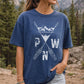 A woman wearing a faded navy blue t-shirt with a white distressed graphic of mountains, crossed arrows, and the letters P, W, and N, set against a blurred forest background.