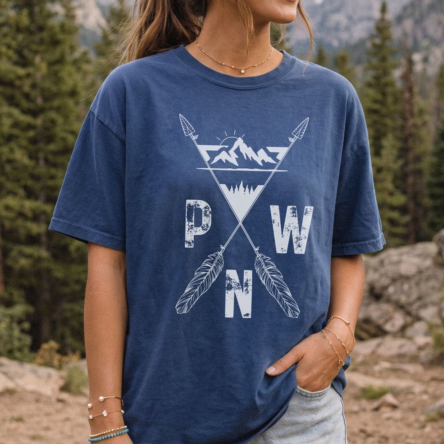 A woman wearing a faded navy blue t-shirt with a white distressed graphic of mountains, crossed arrows, and the letters P, W, and N, set against a blurred forest background.