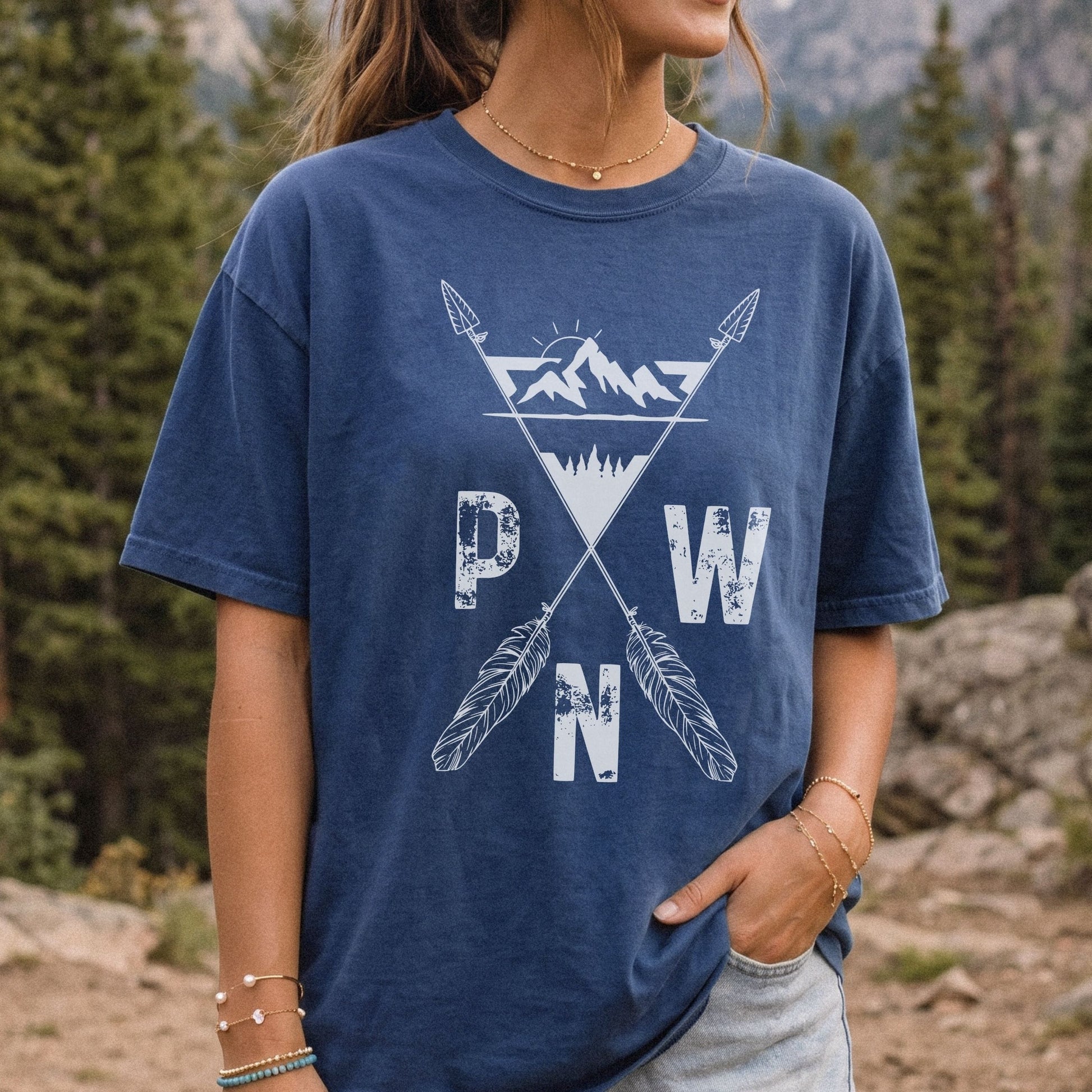 A woman wearing a faded navy blue t-shirt with a white distressed graphic of mountains, crossed arrows, and the letters P, W, and N, set against a blurred forest background.