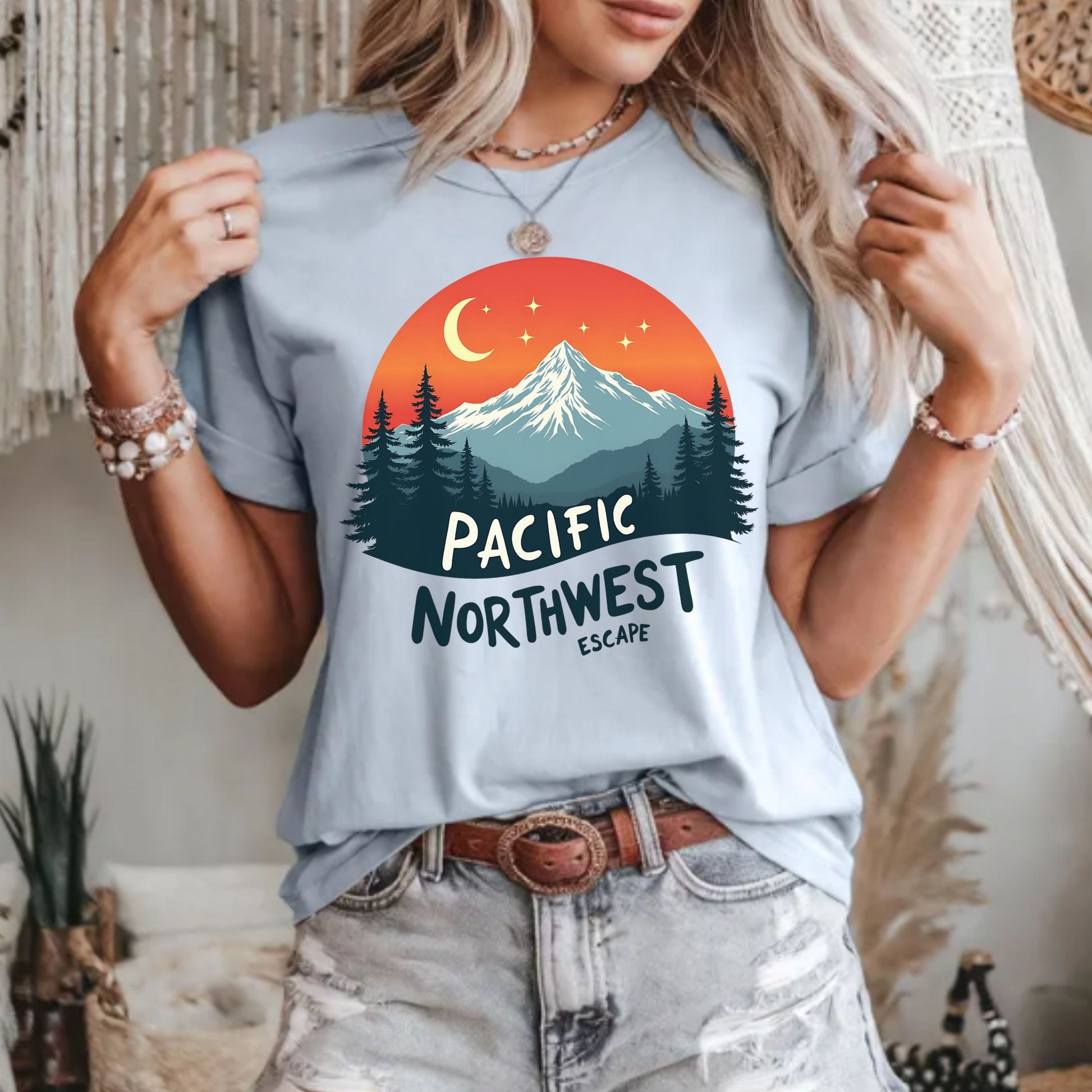 A woman wearing a light blue graphic t-shirt with a 'Pacific Northwest Escape' mountain and sunset design, paired with distressed light-wash jeans.