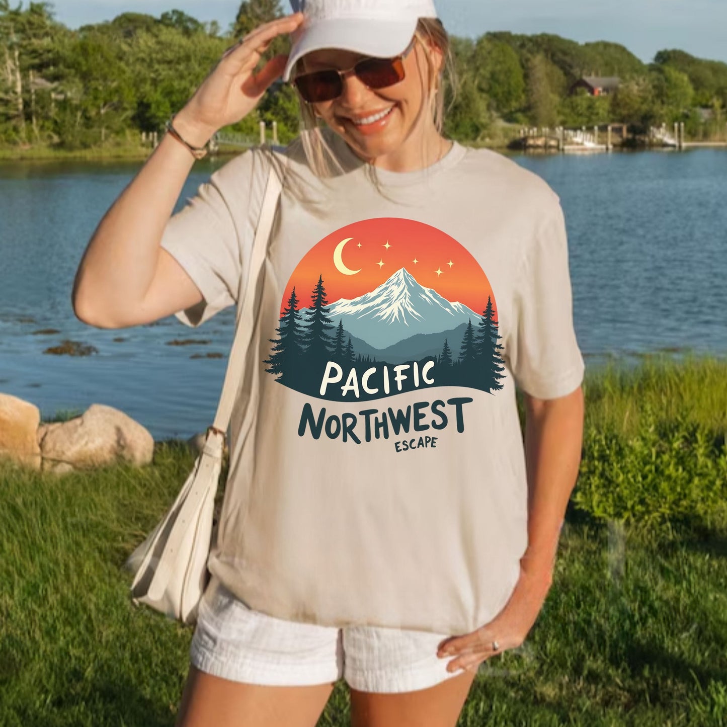 A woman wearing a beige graphic t-shirt with a 'Pacific Northwest Escape' mountain and moon design, standing outdoors near a lake with trees in the background.