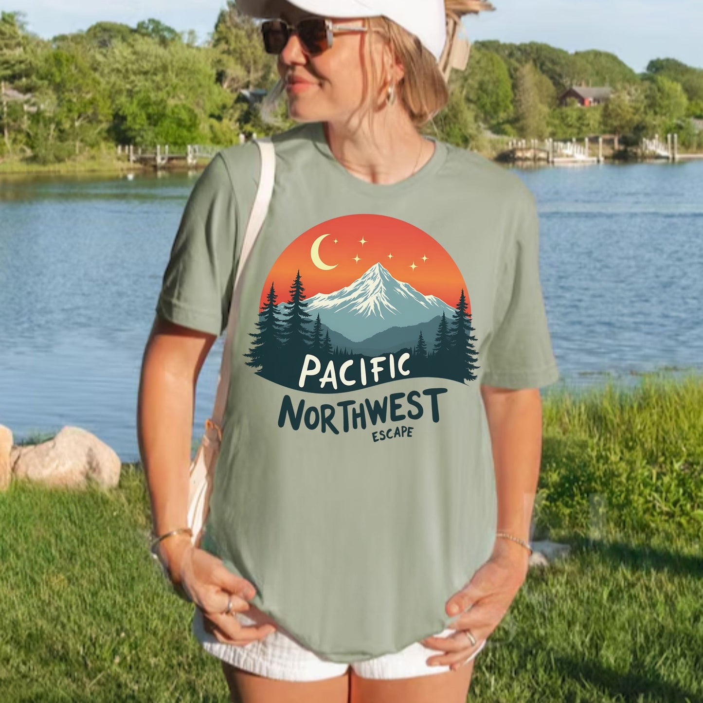 A woman wearing a sage green graphic t-shirt with a 'Pacific Northwest Escape' mountain sunset print, standing outdoors by a lake.