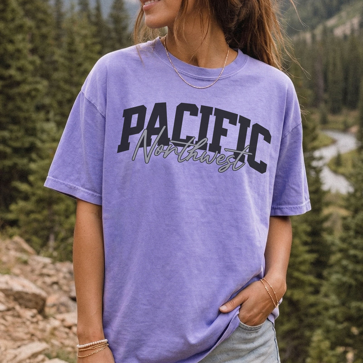 A woman wearing a lavender-purple t-shirt with 'PACIFIC Northwest' printed across the chest, set against a blurred background of a forest and mountain landscape.