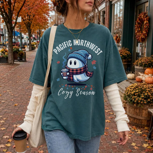 A woman standing on a brick street wearing a teal graphic T-shirt with a cartoon ghost in a blue beanie and plaid scarf holding a coffee cup, with the text 'PACIFIC NORTHWEST Cozy Season.' She has a cream long-sleeve shirt underneath, blue jeans, and a cream shoulder bag.