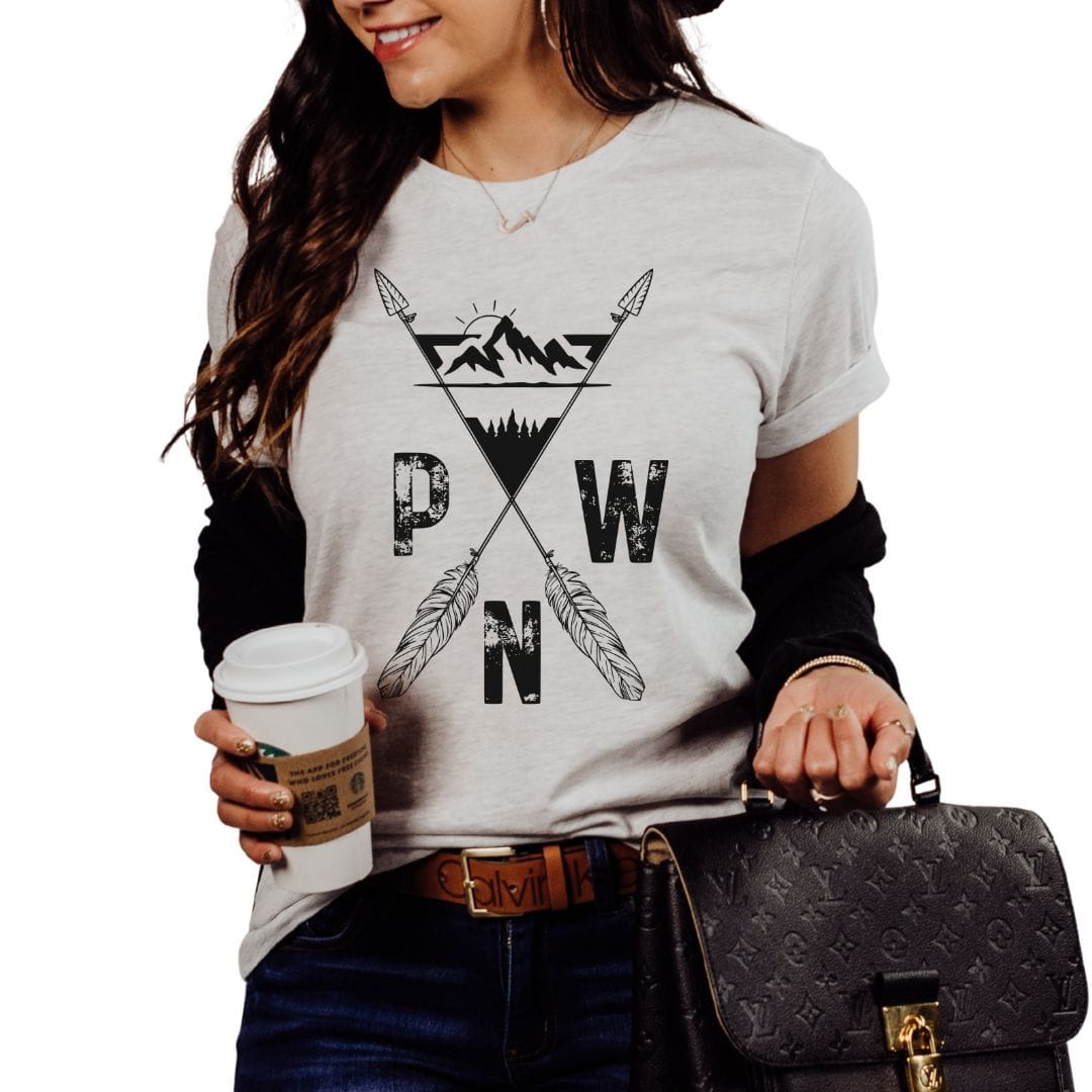 A woman wearing a light gray heather t-shirt with a black graphic of crossed arrows, mountains, and the letters 'PWN'. She is holding a coffee cup and a black monogram handbag against a plain white background.