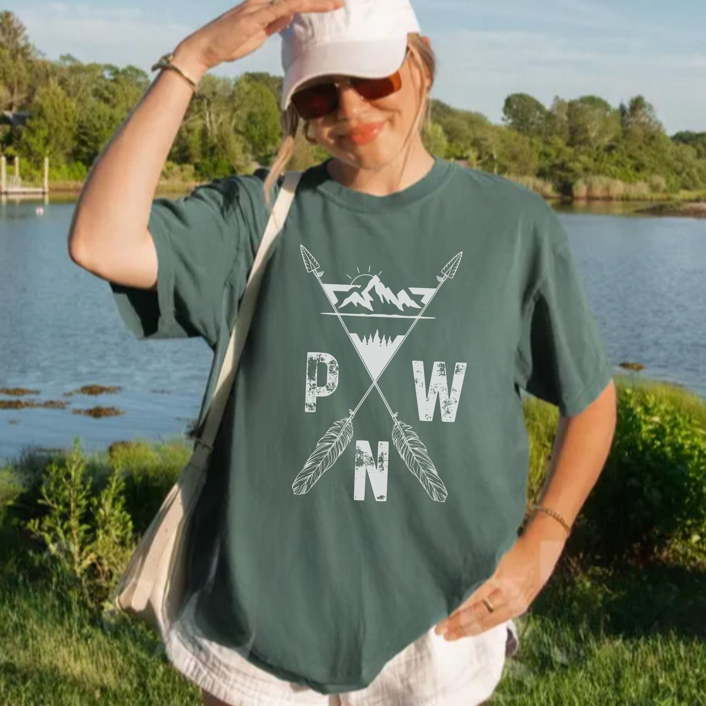 A woman outdoors by a lake wearing an oversized dark green T-shirt with a white graphic print featuring crossed arrows, mountains, and the letters P, W, and N. She is also wearing a white cap, sunglasses, and a light-colored shoulder bag.