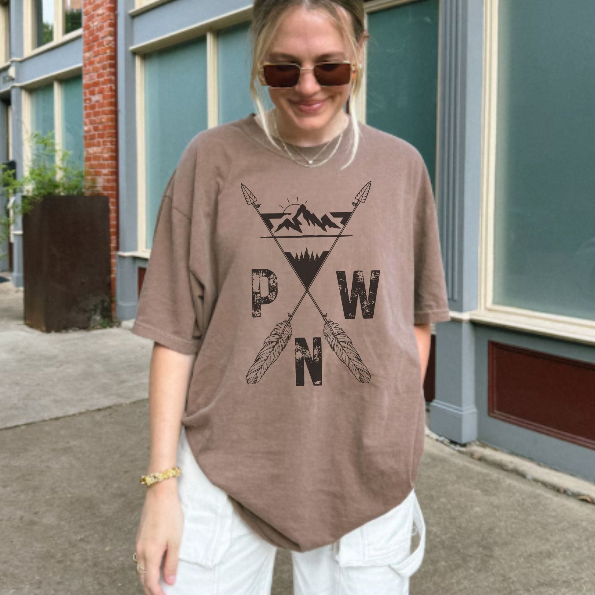 A woman wearing an oversizedespresso brown t-shirt with a black graphic of mountains, crossed arrows, and the letters 'PWN', paired with white cargo pants, standing outdoors in front of a building.