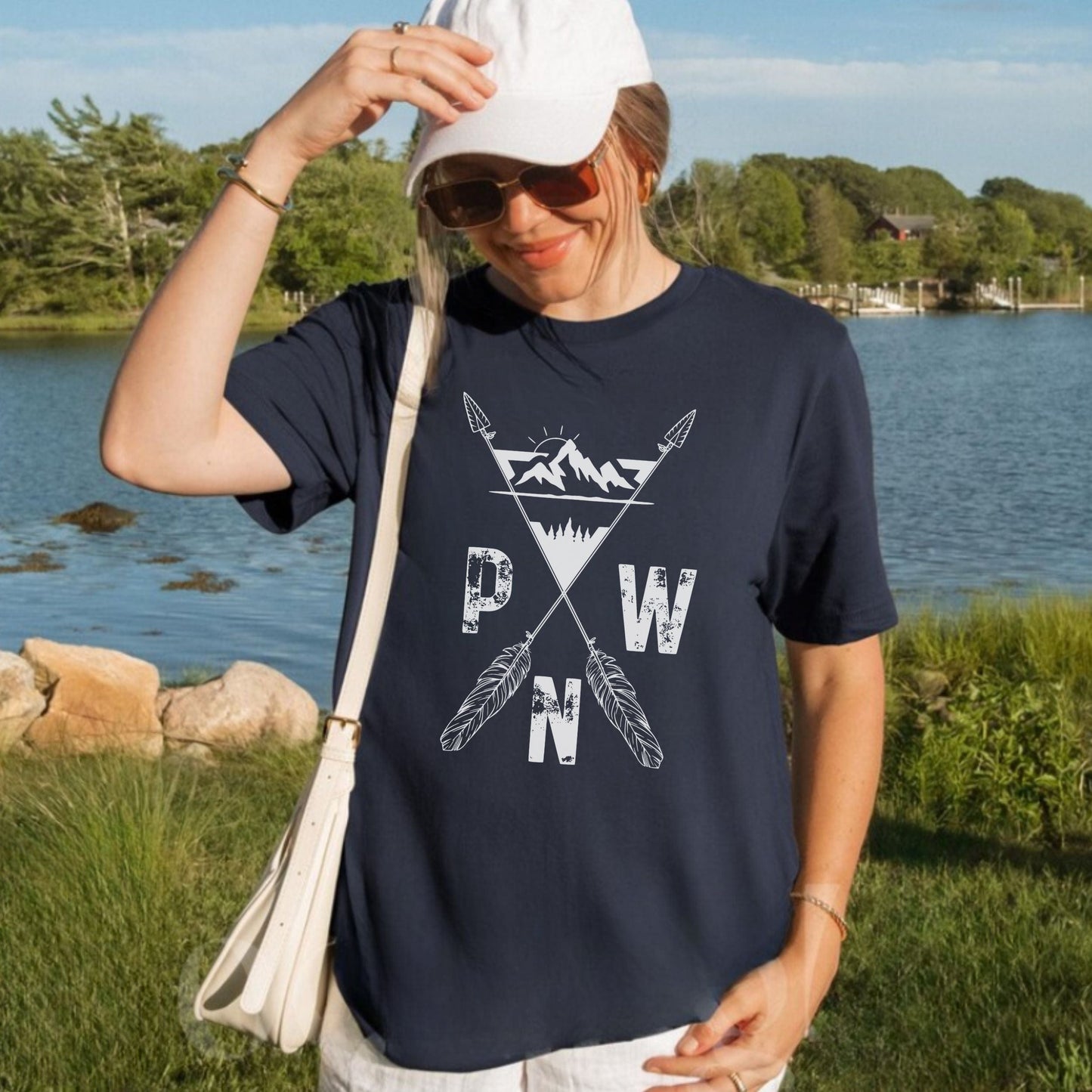 Person wearing a navy blue t-shirt with a white PNW mountains and arrows, graphic design outdoors by a lake.