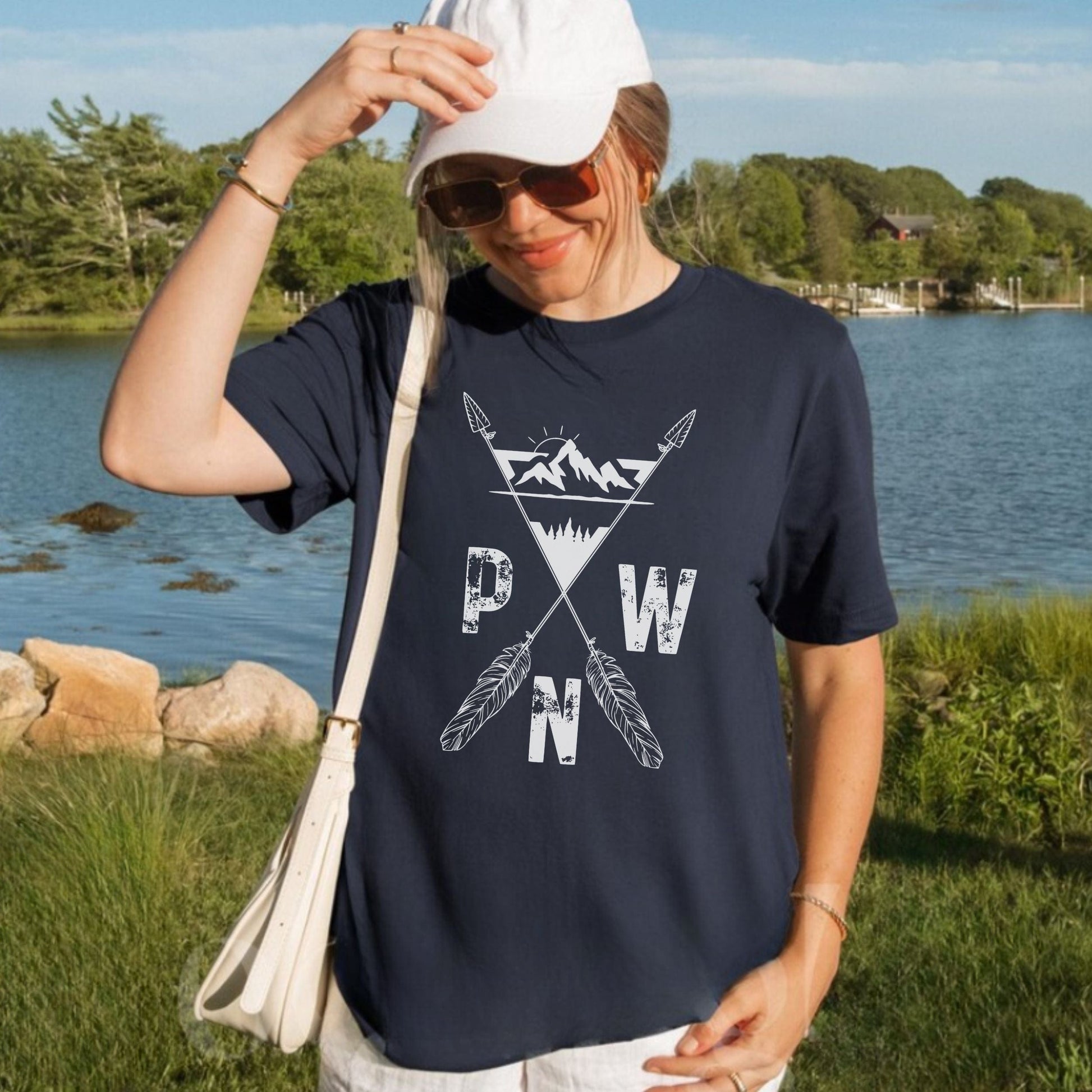 Person wearing a navy blue t-shirt with a white PNW mountains and arrows, graphic design outdoors by a lake.