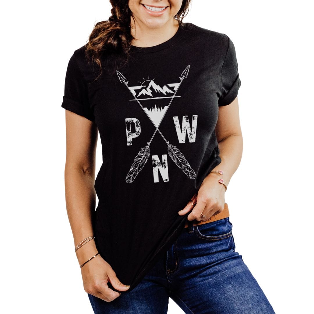 A woman wearing a black short-sleeve graphic T-shirt with a white distressed mountain, crossed arrows, and the letters P, W, and N on the front, paired with blue jeans against a plain white background.