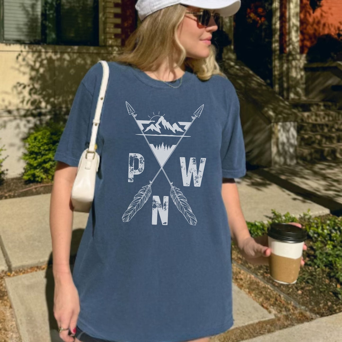 A woman wearing a blue graphic t-shirt with a white mountain and crossed arrows design featuring the letters 'PWN', walking outdoors on a sidewalk while holding a coffee cup.