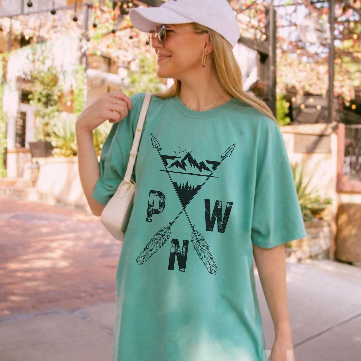 A woman wearing a light green graphic t-shirt with a black 'PWN' mountain and feather design, styled with a white cap, sunglasses, and a white shoulder bag in an outdoor setting.