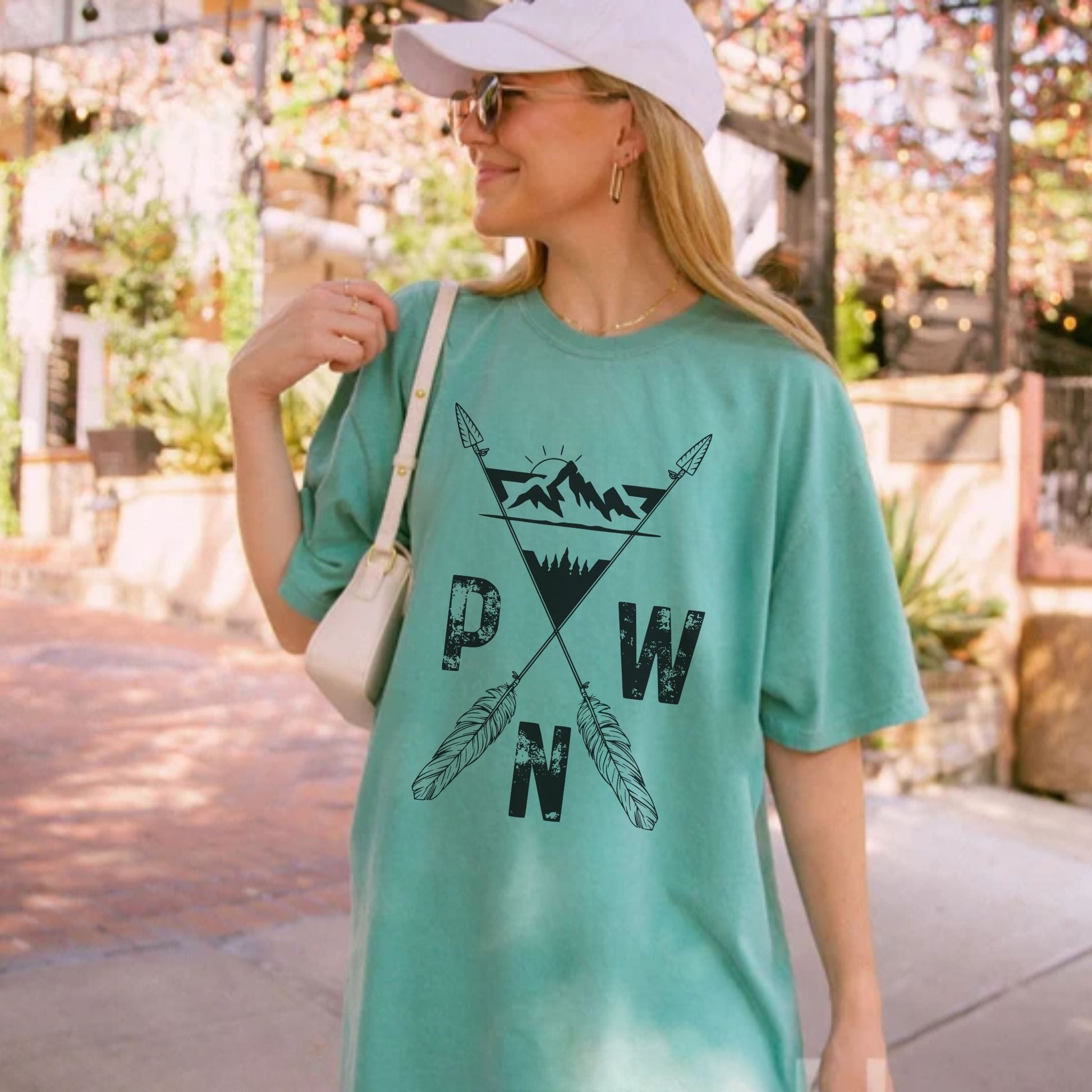 A woman wearing a light green graphic t-shirt with a black 'PWN' mountain and feather design, styled with a white cap, sunglasses, and a white shoulder bag in an outdoor setting.