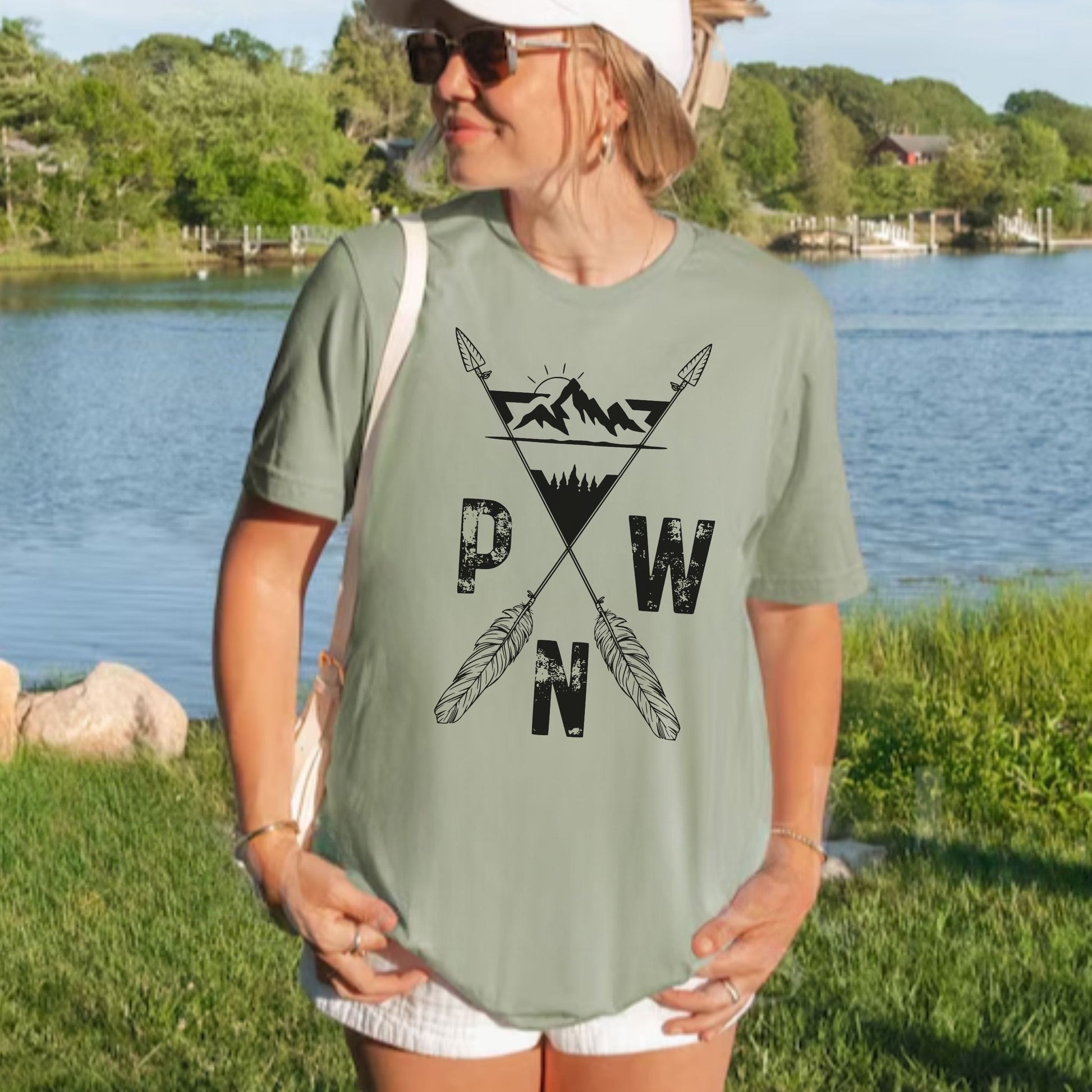 A woman wearing a sage green t-shirt with a black 'PWN' mountain and feather graphic, standing outdoors near a lake with trees in the background.