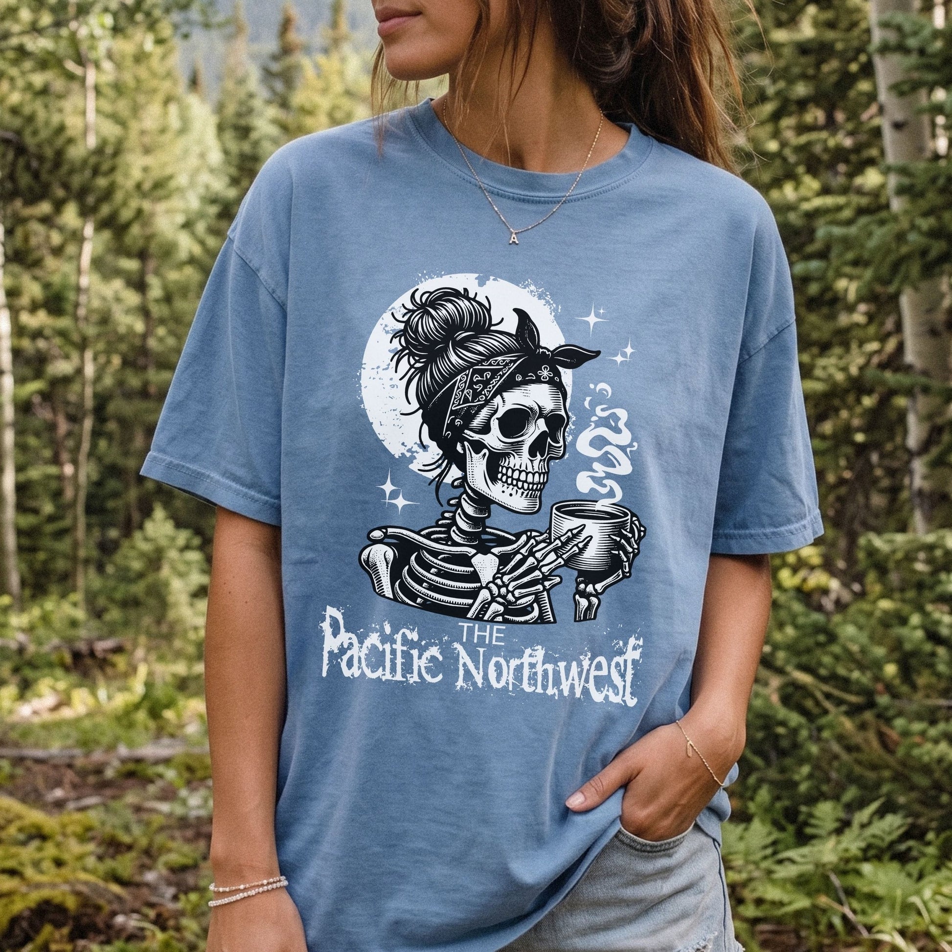 A woman wearing a light blue graphic t-shirt in an outdoor forest setting. The shirt features a black and white illustration of a skeleton holding a mug in front of a moon, with the text 'THE Pacific Northwest' printed below.