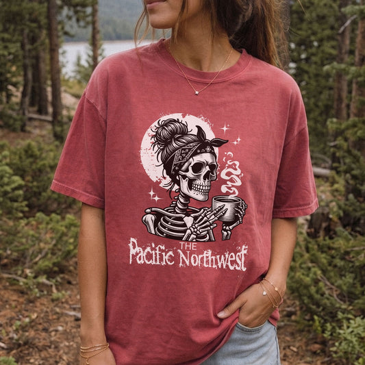 A woman wearing a faded dusty rose graphic t-shirt with a skeleton holding a mug and the text 'The Pacific Northwest' in front of a forest background.