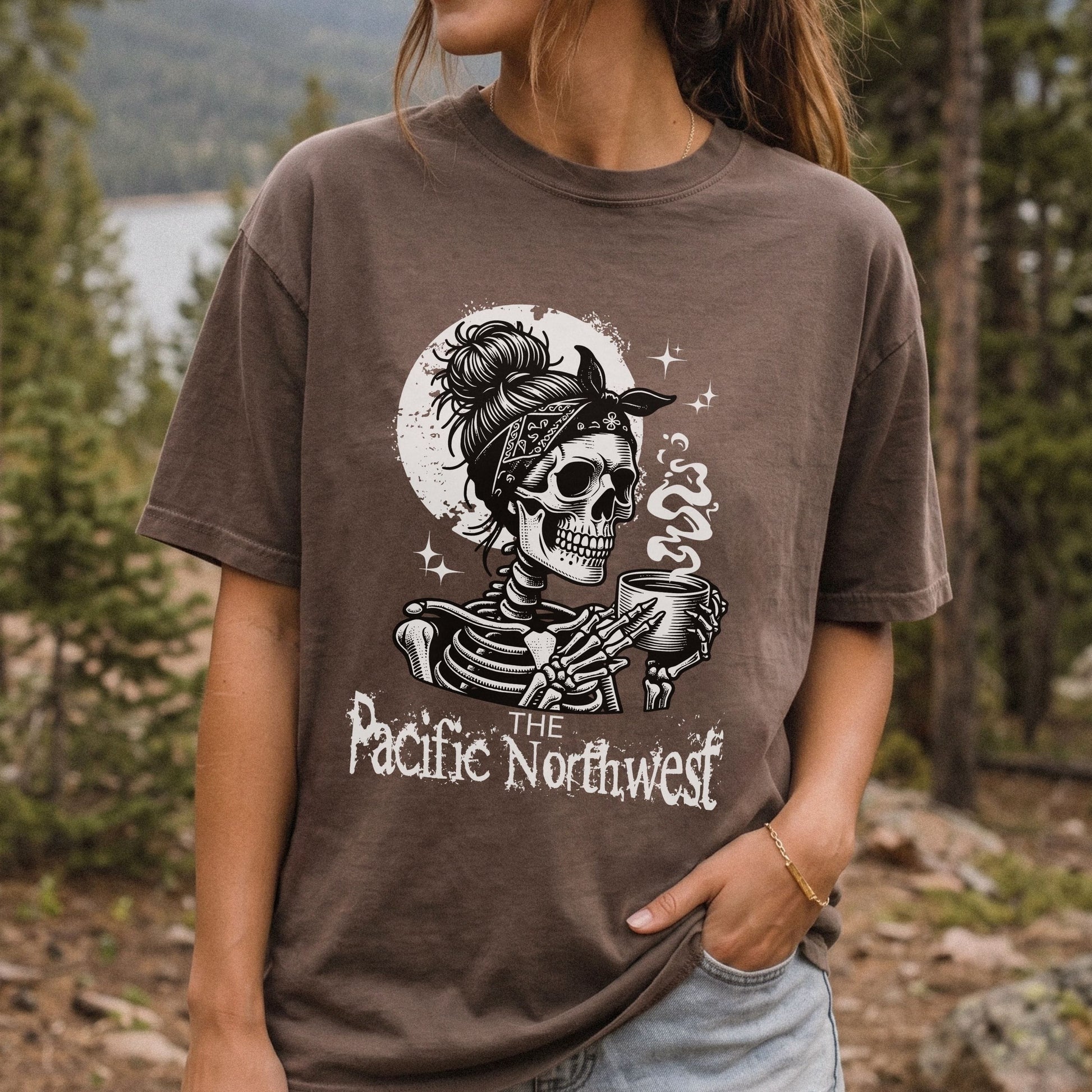 A woman wearing a brown graphic t-shirt with a black and white design of a skeleton holding a mug, with the text 'THE Pacific Northwest' on the front, set against a blurred forest background.