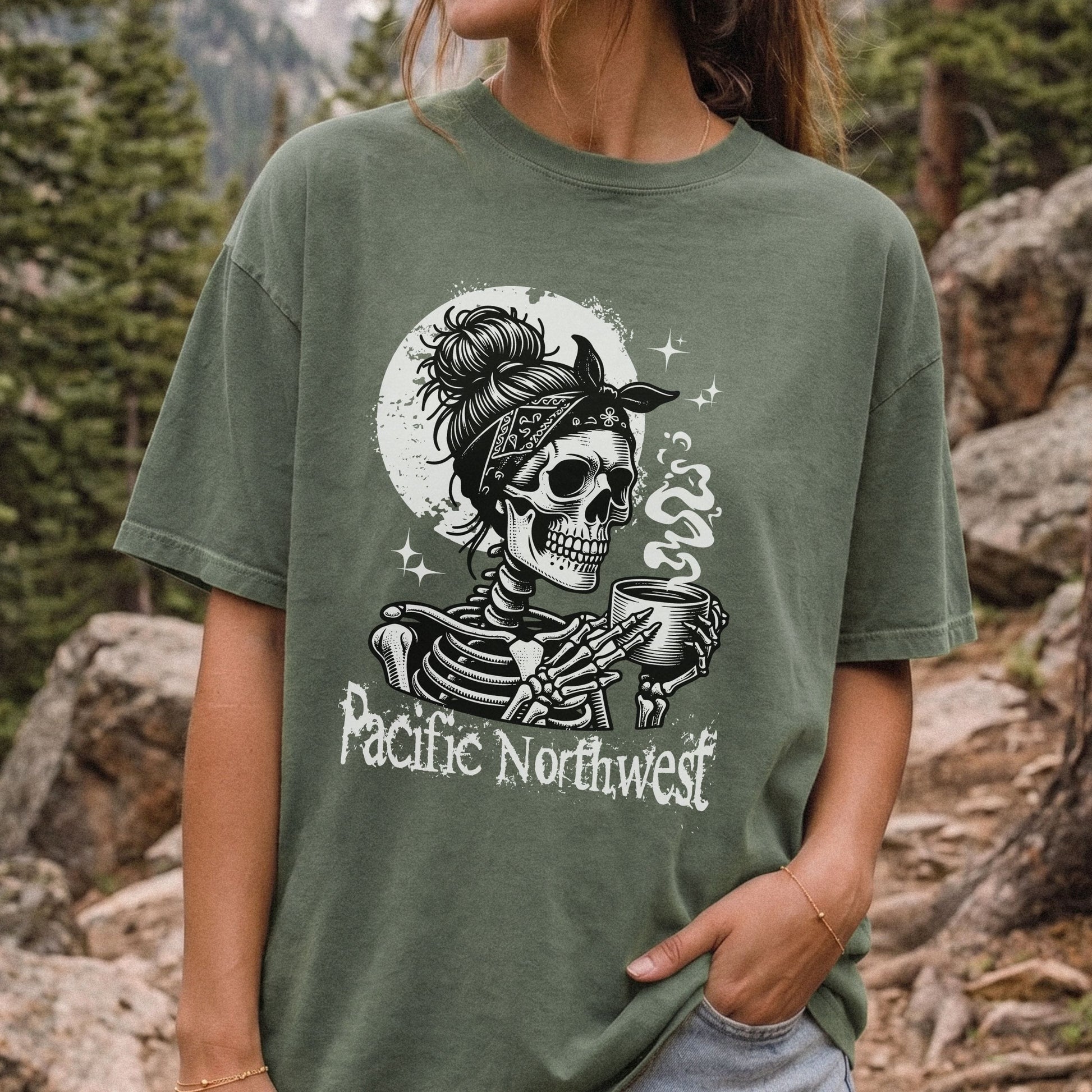 A woman wearing an oversized moss green graphic t-shirt in an outdoor mountain setting. The t-shirt features a black and white graphic of a skeleton wearing a bandana and holding a mug, with the text 'Pacific Northwest' printed below.