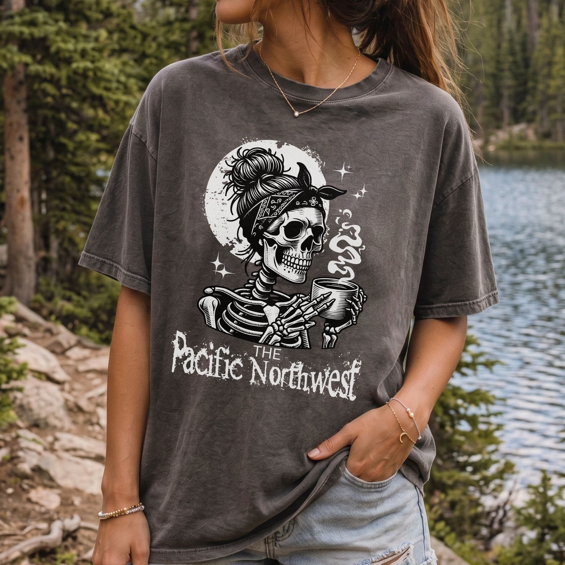 A woman wearing a dark gray mineral-wash graphic t-shirt with a skeleton holding a mug and the text 'The Pacific Northwest' in front of a forest and lake background.