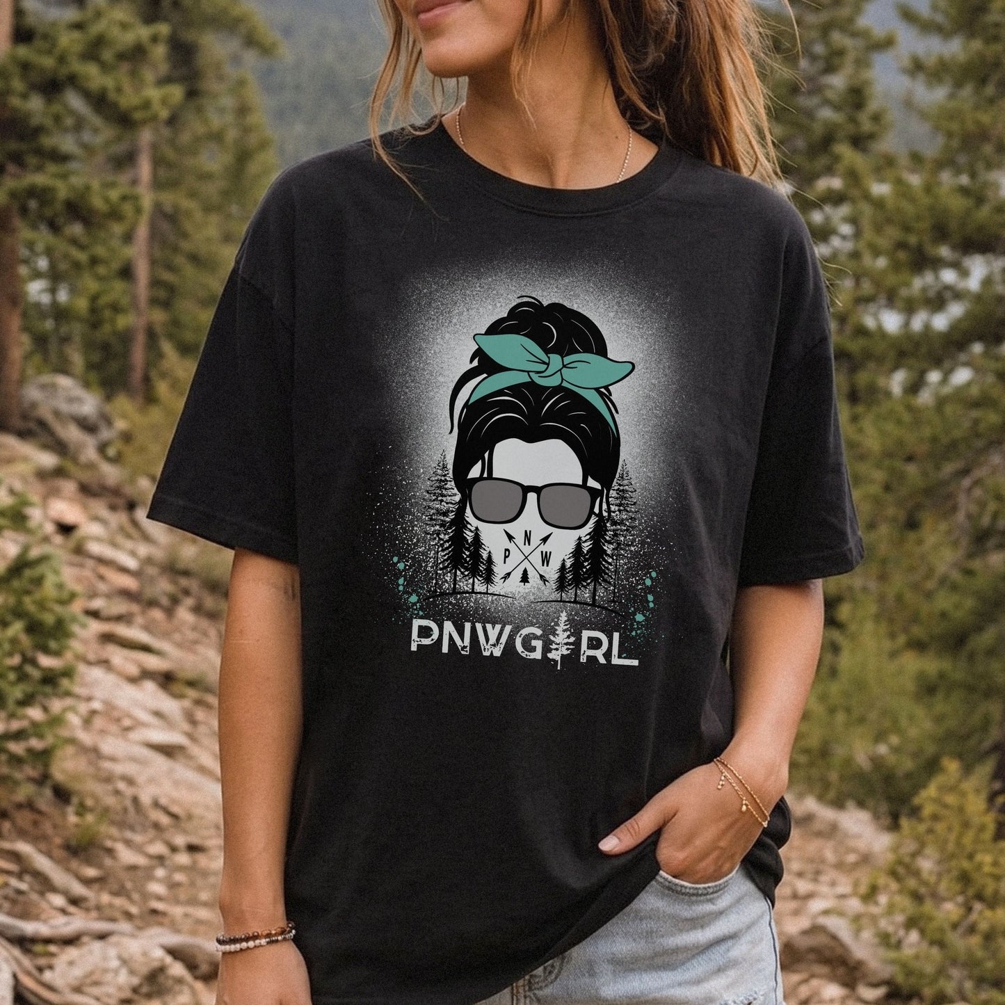 A woman wearing a black graphic t-shirt with a 'PNW GIRL' skull and bandana design, photographed outdoors in a forest setting.