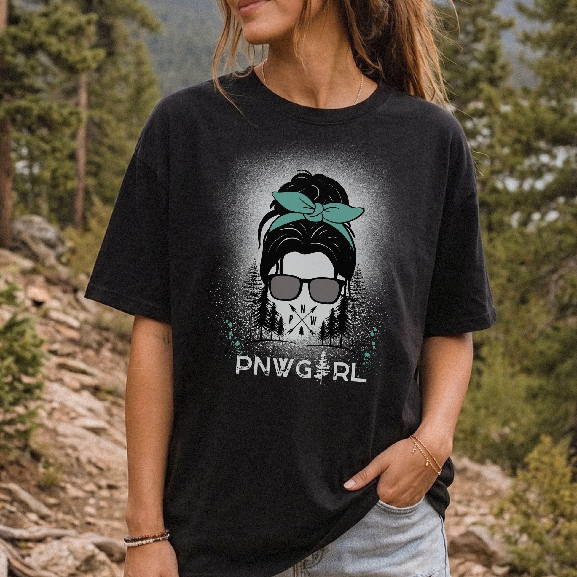 A woman wearing a black graphic t-shirt with a 'PNW GIRL' skull and bandana design, photographed outdoors in a forest setting.