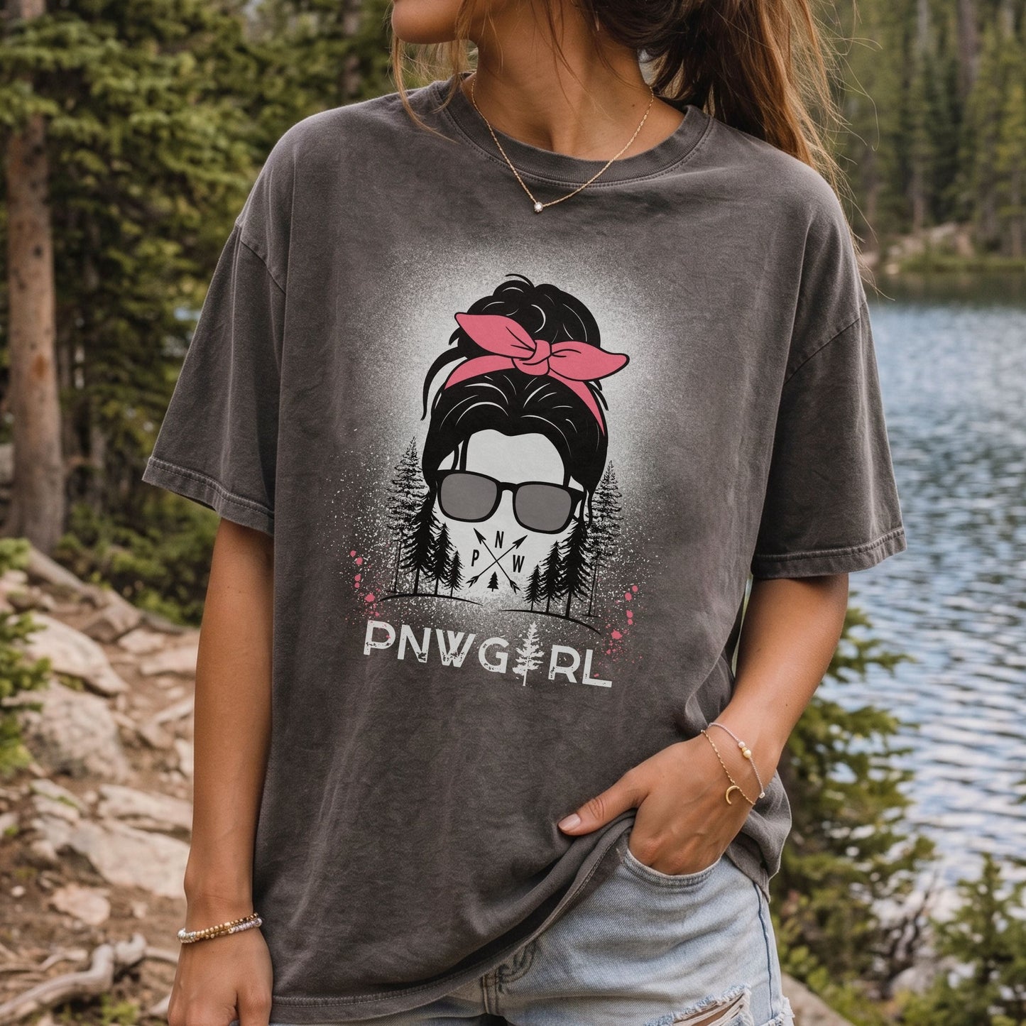 A woman wearing a faded charcoal gray graphic t-shirt with a 'PNW Girl' design, standing outdoors near a lake and pine trees.
