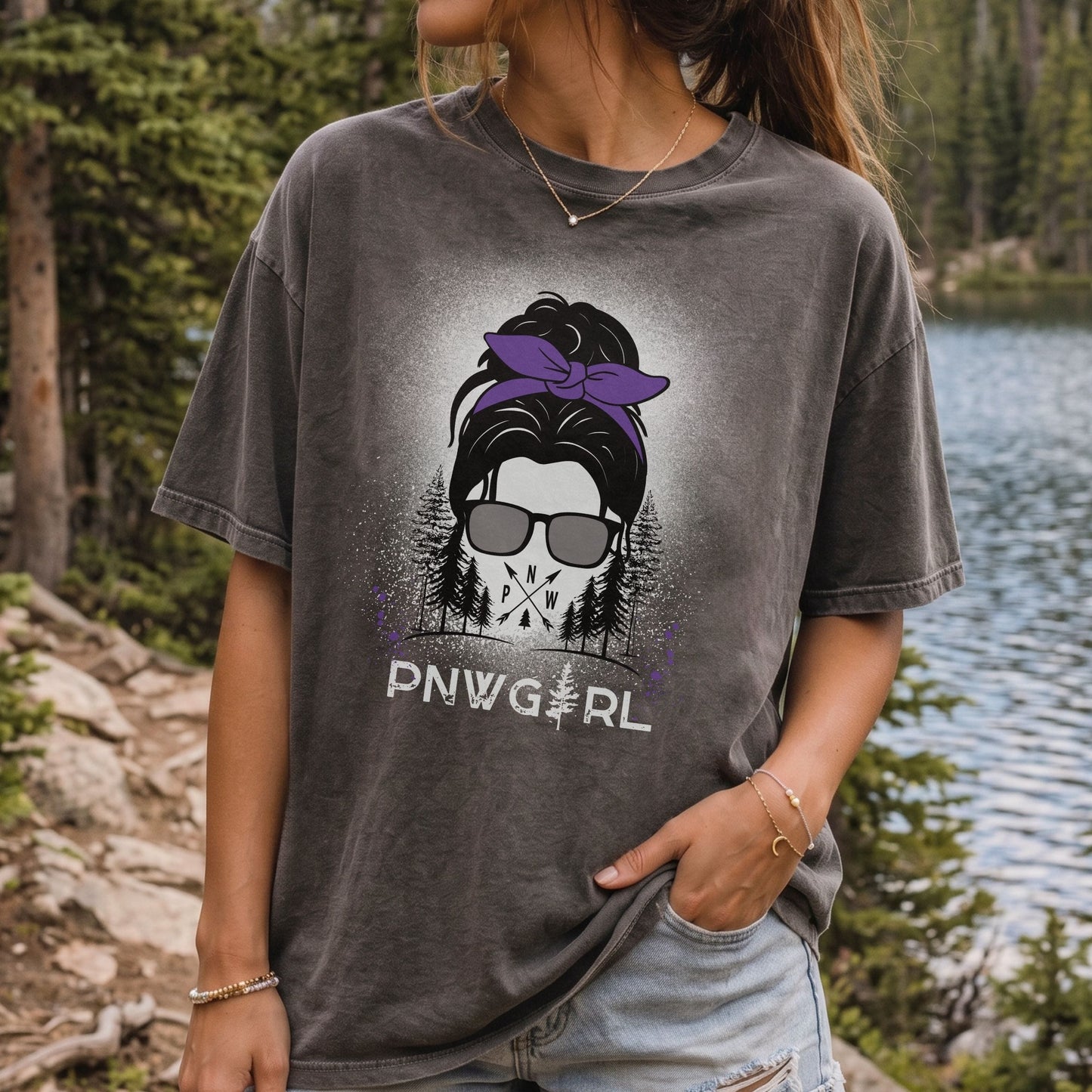 A woman wearing a faded charcoal gray graphic t-shirt with a purple and black design and the text 'PNW GIRL' printed on the front, standing outdoors near a lake and pine trees.