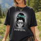 A woman wearing a dark charcoal graphic t-shirt with a 'PNWG GIRL' design, styled outdoors in a forest setting with an olive green cap, plaid shirt tied around her waist, and shorts.